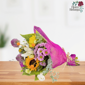 Ramo primaveral con girasol, alium, margaritas, lisianthum, craspedia y eucalipto, envuelto en papel rosa sobre mesa de madera.