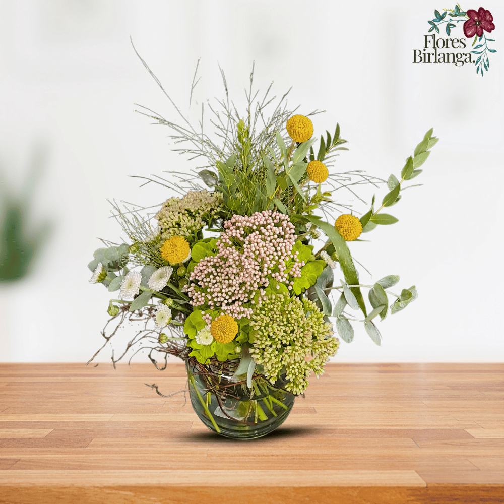 Arreglo floral en jarrón de cristal con lisianthus rosado, rosas blancas y verdes, statice morado y follaje sobre mesa de madera.