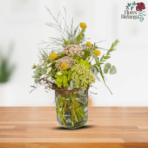 Arreglo floral en jarrón de cristal con flor de cera, sedum, eucalipto, hortensia, margarita de botón y esparraguera sobre mesa de madera.