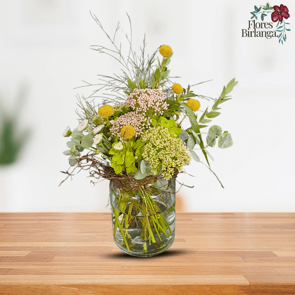 Arreglo floral en jarrón de cristal con flor de cera, sedum, eucalipto, hortensia, margarita de botón y esparraguera sobre mesa de madera.