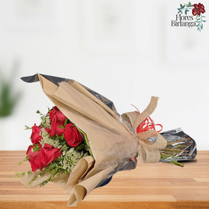 Ramo de 6 rosas rojas con brunia, flor de arroz, aster y flor de cera, envuelto en celofán transparente y decorado con cintas de rafia, sobre mesa de madera.