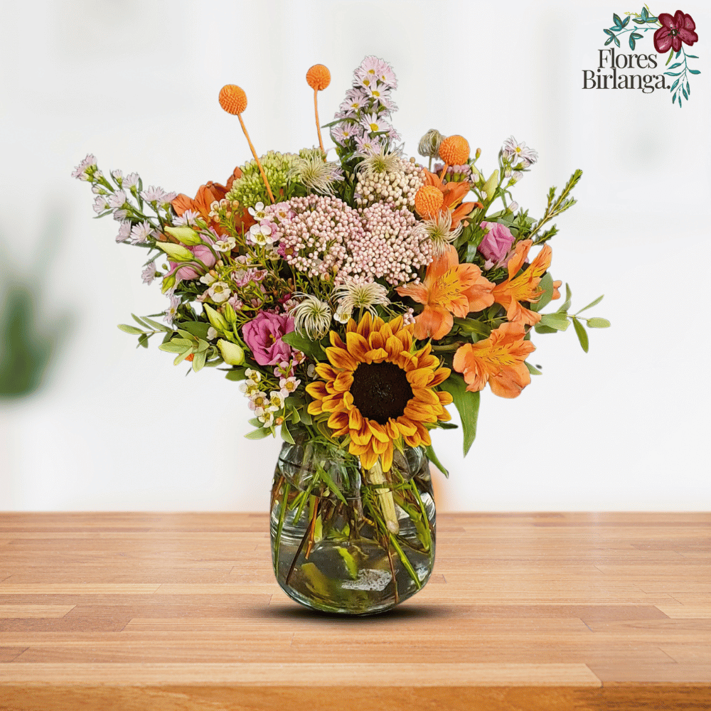 Arreglo floral en jarrón de cristal con girasol, rosas, alstroemeria, lisianthus, craspedia y follaje verde sobre mesa de madera.
