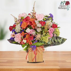 Arreglo floral con claveles rosados, rosas rojas y naranjas, statice morado, achillea blanca y follaje verde en base de papel sobre mesa de madera.