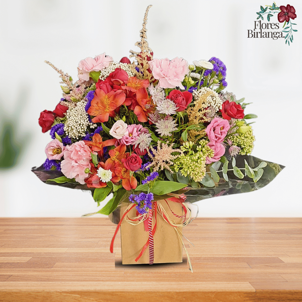 Arreglo floral con claveles rosados, rosas rojas y naranjas, statice morado, achillea blanca y follaje verde en base de papel sobre mesa de madera.