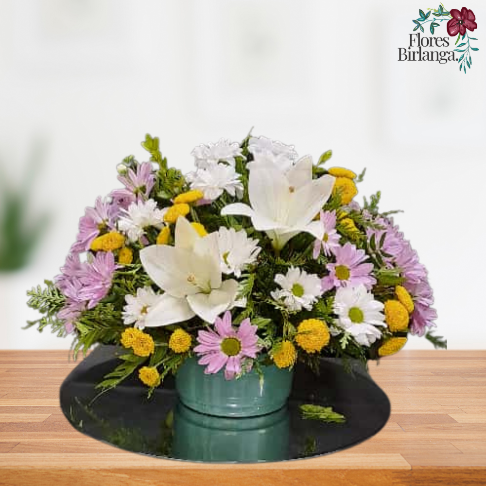 Centro floral redondo y frondoso para Todos los Santos con lirios blancos, margaritas blancas, crisantemos morados y flores de relleno amarillas, sobre una mesa de madera.